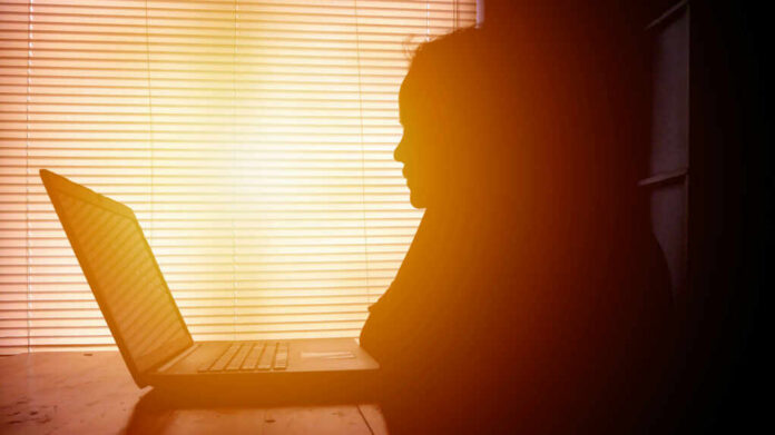 Silhouette of a person working on a laptop with sunlight streaming through blinds