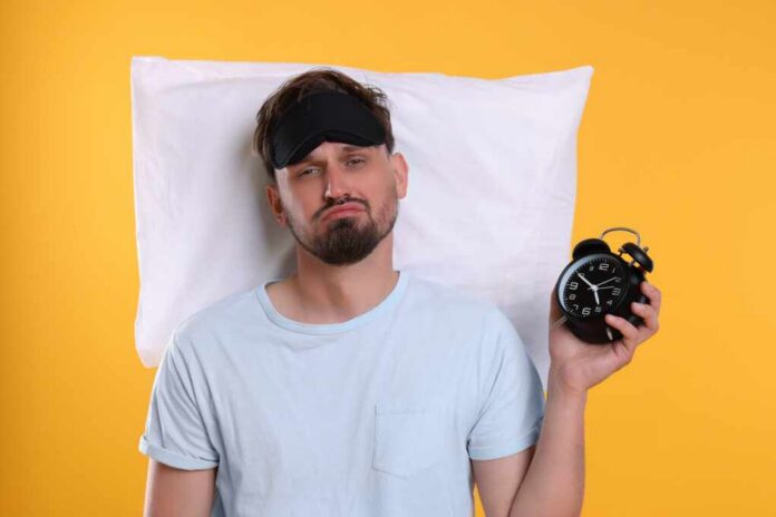 shutterstock_2361618683.jpg Man wearing a sleep mask holding an alarm clock with a frustrated expression
