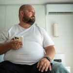 Couch Potato Lifestyle Linked to Dementia A man in a white t-shirt sitting on a bed, holding a smartphone and looking thoughtfully out of a window