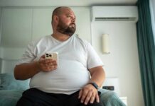 A man in a white t-shirt sitting on a bed, holding a smartphone and looking thoughtfully out of a window