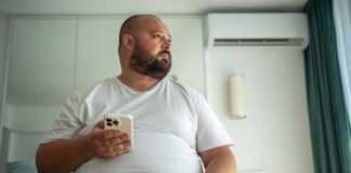A man in a white t-shirt sitting on a bed, holding a smartphone and looking thoughtfully out of a window