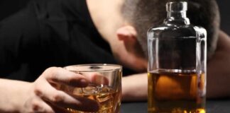 A person with their head down, holding a glass of whiskey next to a bottle