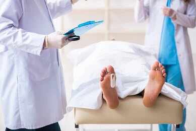 1783083806 Medical staff stand near a covered body on a gurney with a toe tag