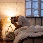 A person sitting on a bed with their head in their hands, expressing distress