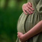 Pregnant woman in a green dress gently holding her belly in a natural setting