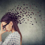 Stress May Be Aging Your Blood A woman with glasses holding her head, with fragments dispersing from her head