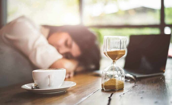 Hourglass on table with coffee cup and sleeping person in background