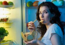 A woman eating a snack while looking at the camera, standing in front of an open refrigerator