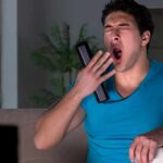 Man yawning on couch holding TV remote and soccer ball
