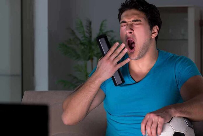 Man yawning on couch holding TV remote and soccer ball