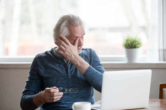 Older man rubbing his eye while sitting at a laptop