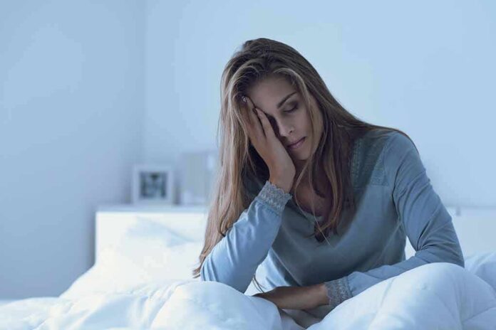 shutterstock_1285836277 Woman sitting in bed with hand on face, looking tired