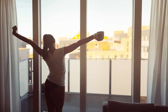 Woman stretching with coffee by a balcony window at sunrise