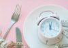 One Meal Daily – Game-Changer Or Health Trap? Pink alarm clock on a white plate with fork, knife, and measuring tape