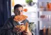 man in robe eating pizza at open refrigerator