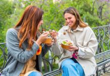 Two women sitting on a park bench eating takeout bowls