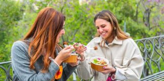 Two women sitting on a park bench eating takeout bowls