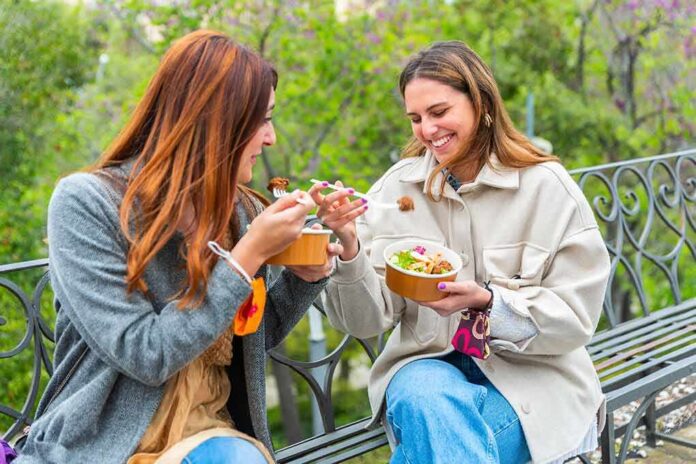 Two women sitting on a park bench eating takeout bowls